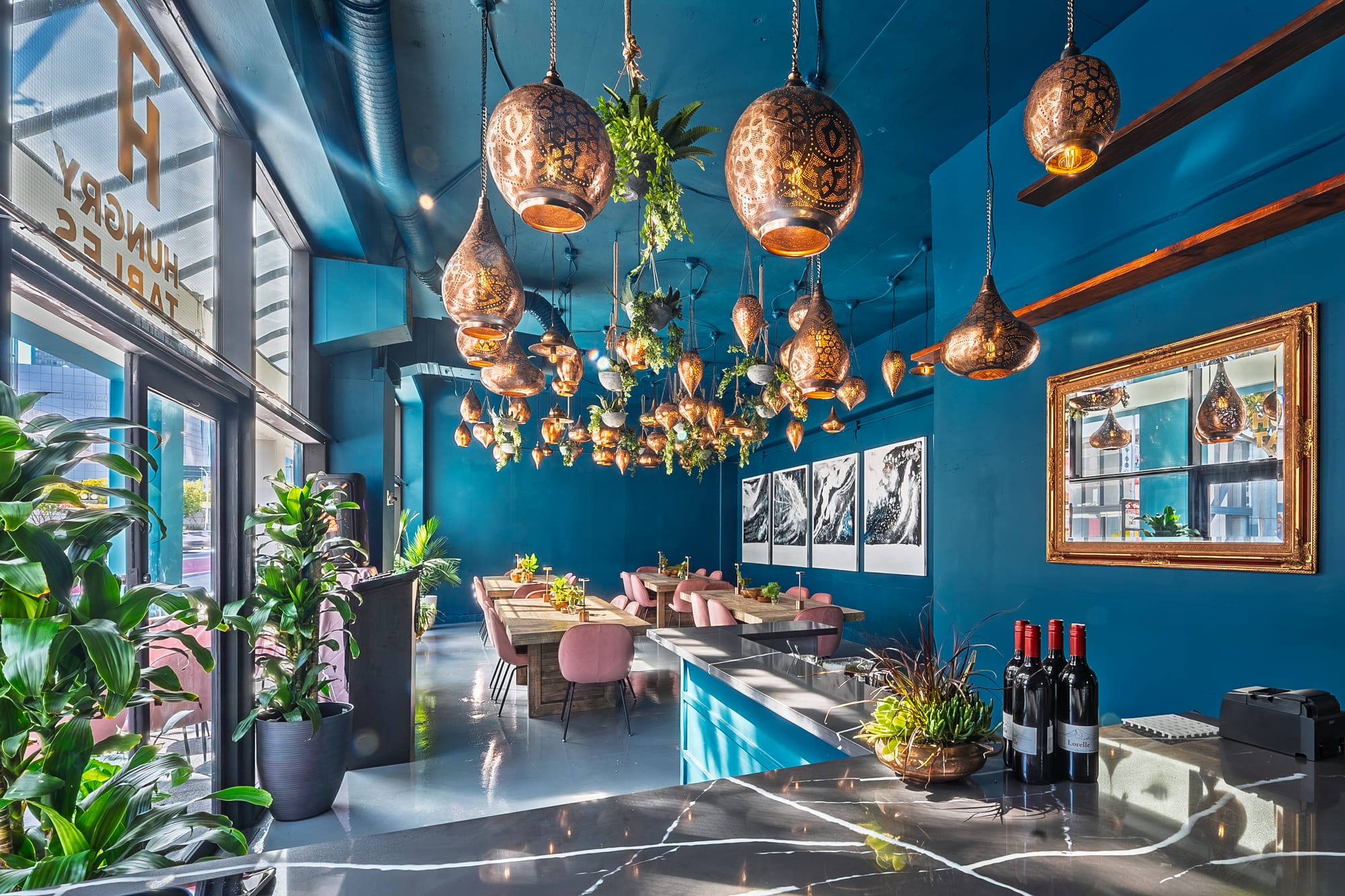 The Hungry Tables bar: teal and marble counter, wine bottles, plants, and copper lanterns overhead