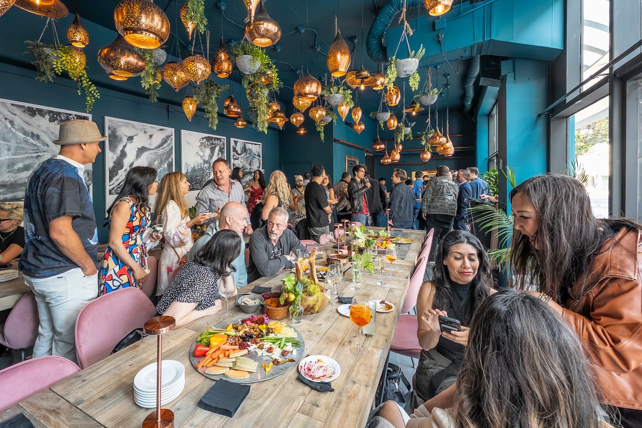 The room at capacity — guests seated the length of the communal table with more standing behind, under the copper lantern ceiling