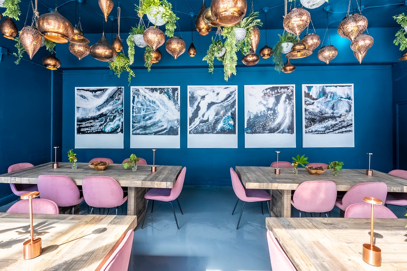 The room at rest: copper lanterns and hanging plants over communal wood tables and rose velvet chairs